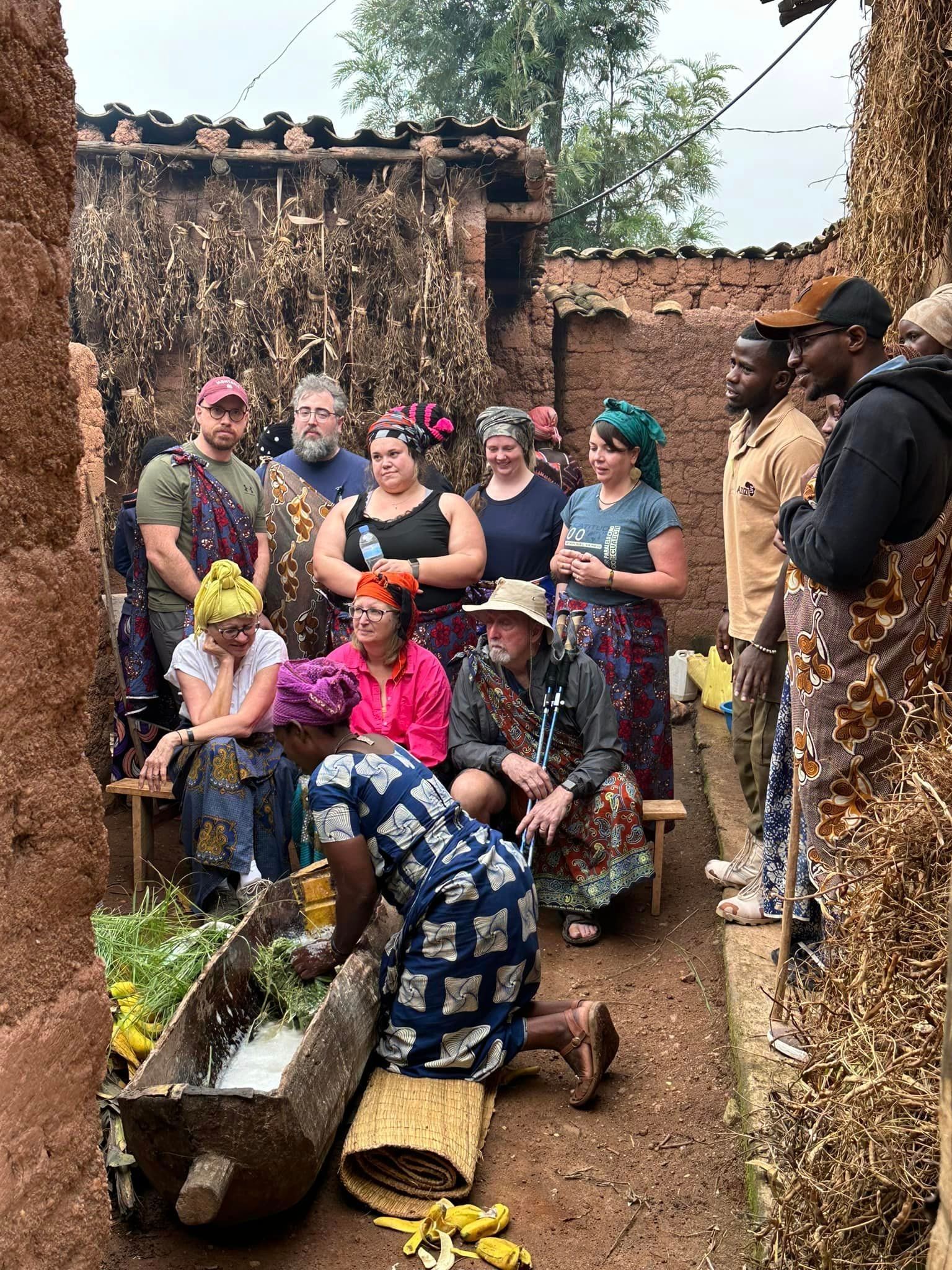 Group of people in African attire, standing and kneeling by a wooden trough. Woman grinds ingredients. Earth-toned buildings.