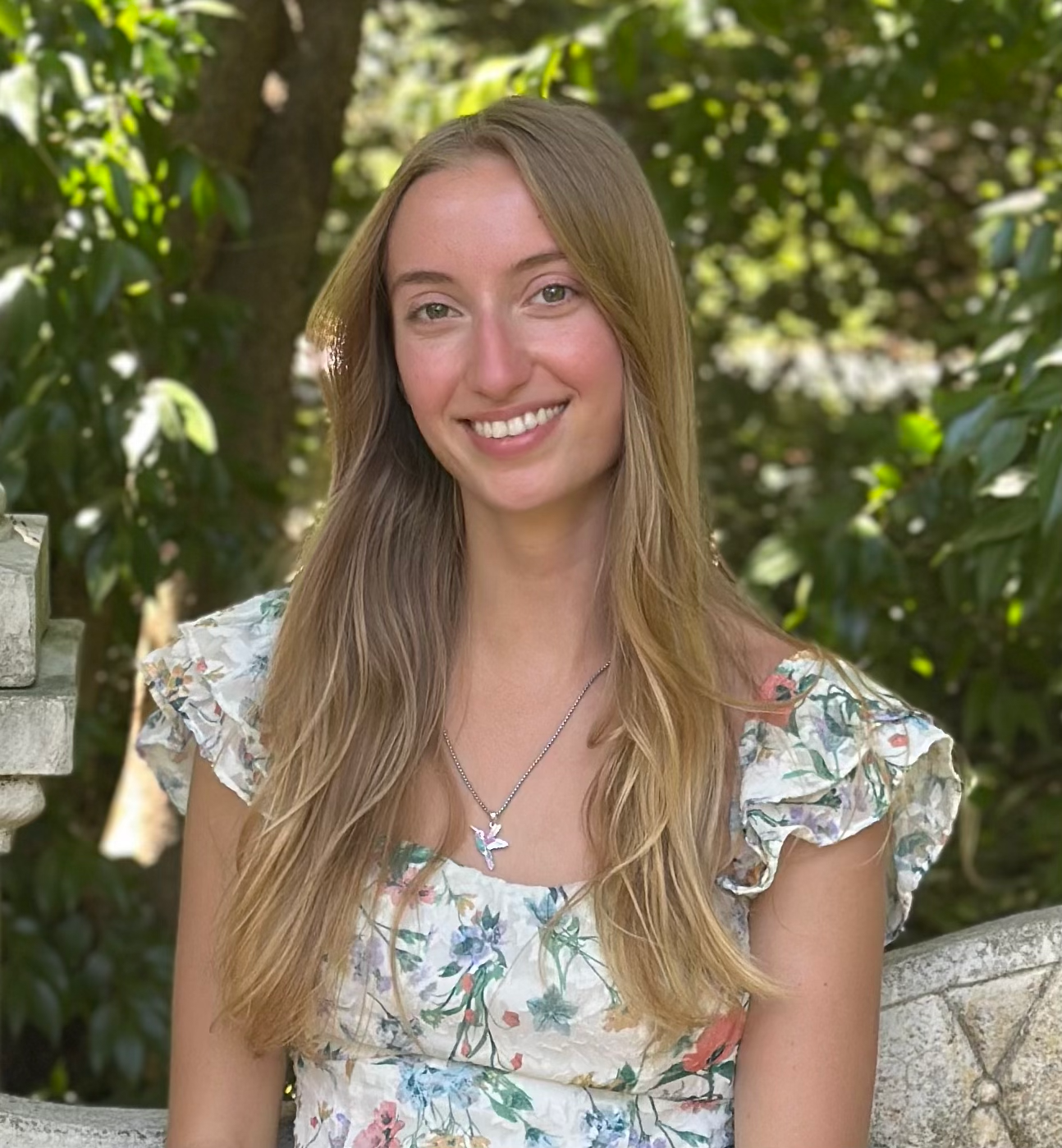 Young woman with long blonde hair smiles outdoors in a floral dress, greenery in background.
