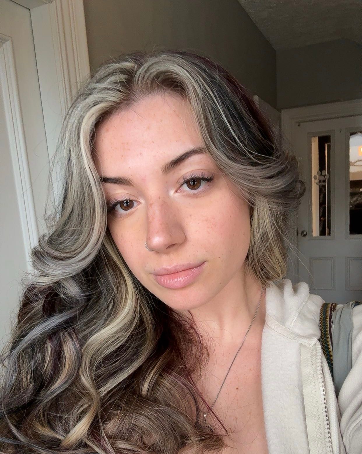 Woman with wavy grey and blonde hair, looking at the camera. She’s wearing a light-colored hoodie indoors.