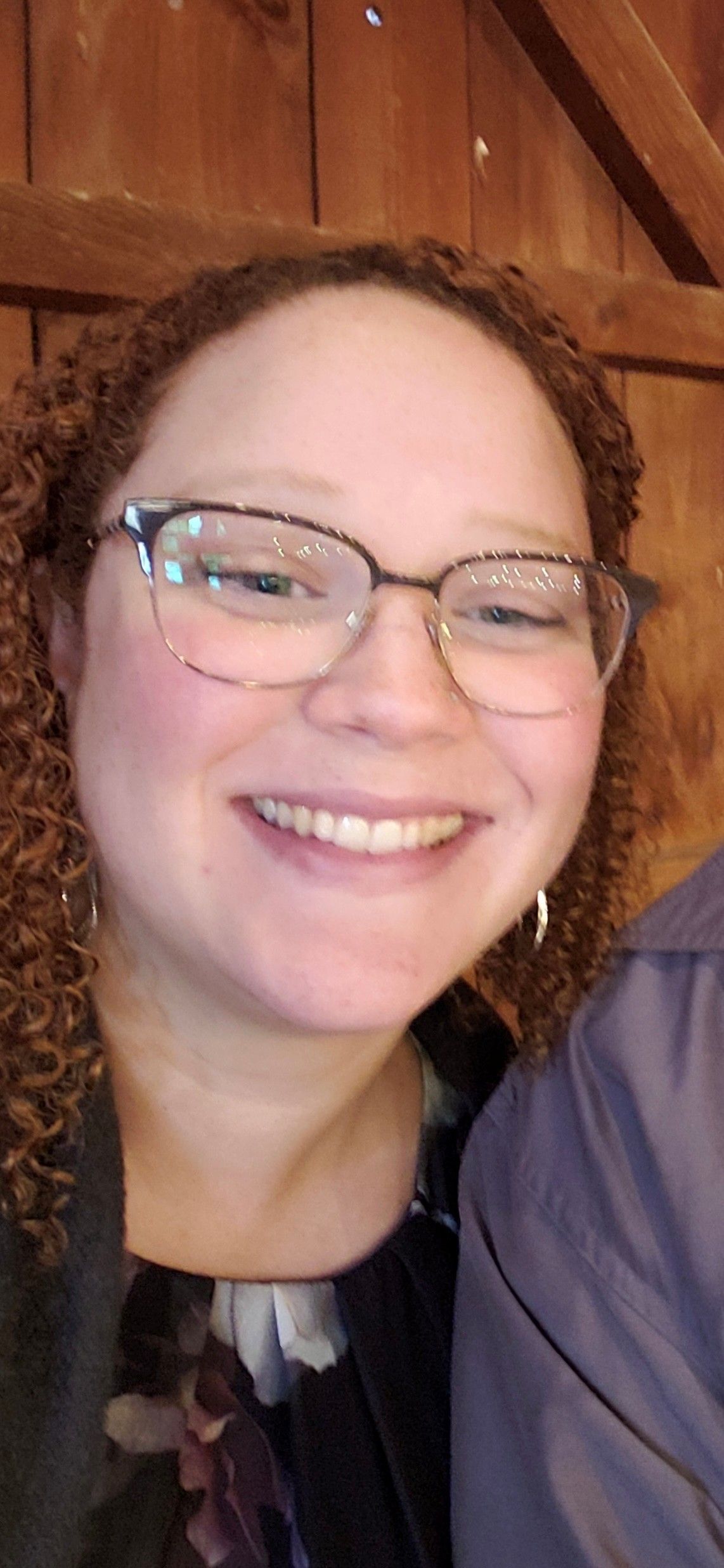 Woman with curly red hair smiling, wearing glasses and a floral top, close-up shot.