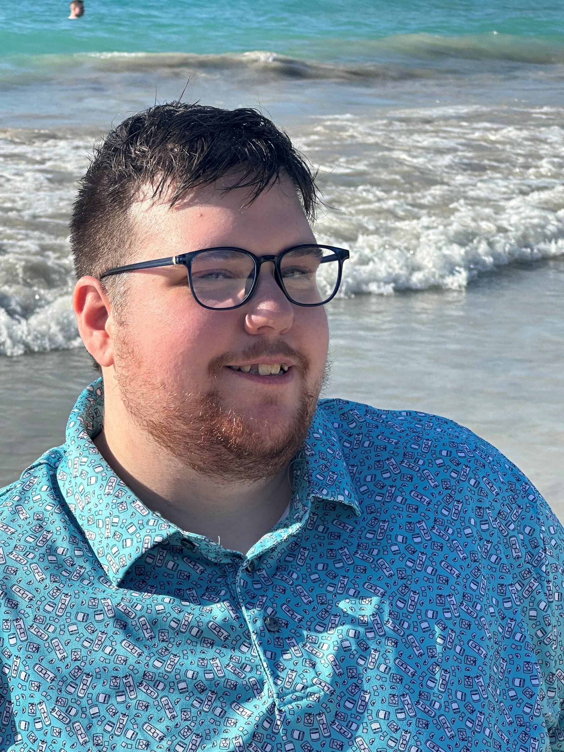 Man with glasses and beard smiles on a beach, wearing a blue floral shirt; ocean in the background.