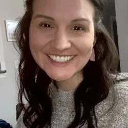 Woman with dark hair smiles; wearing a gray sweater and pink earrings.