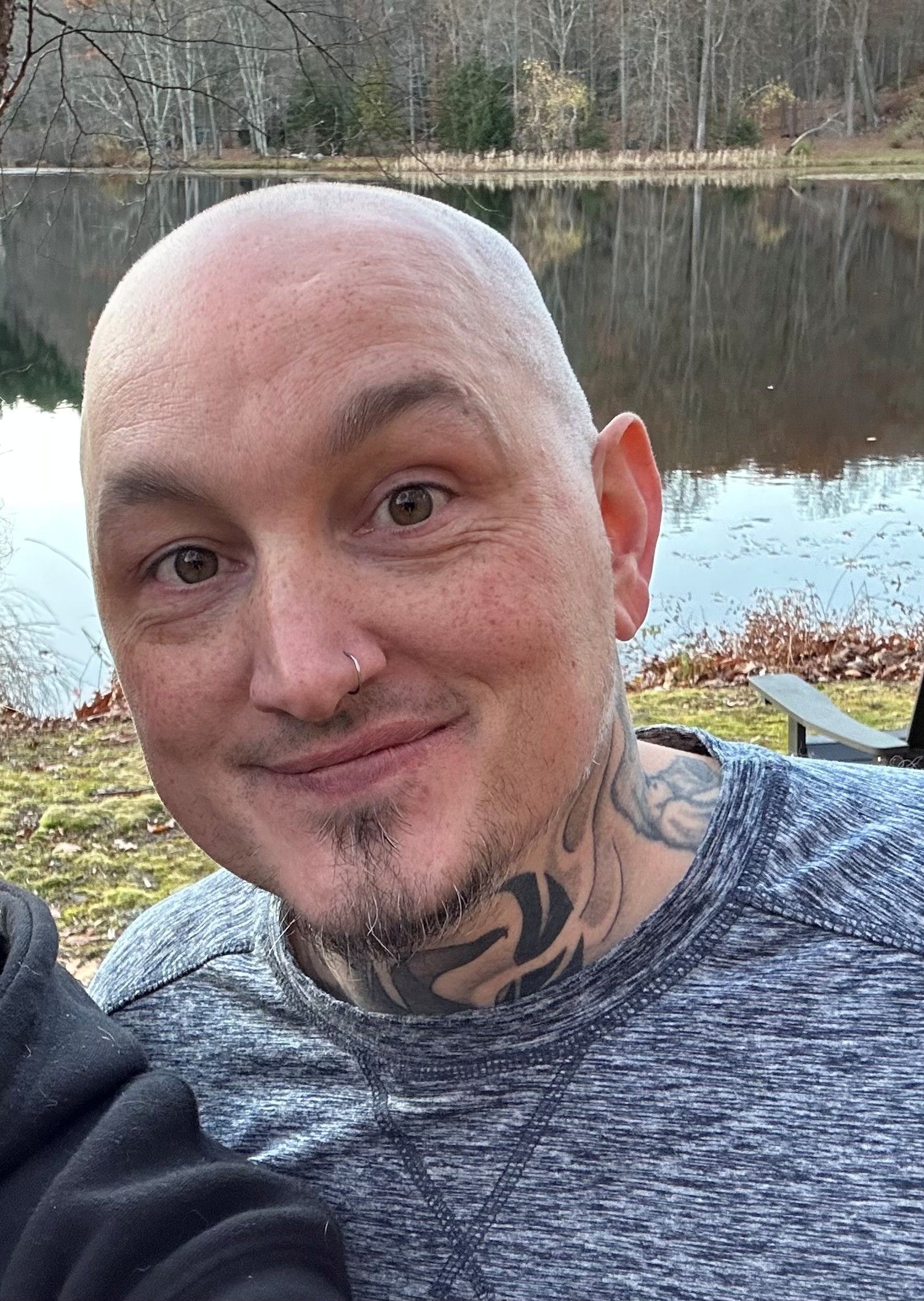 Bald man with neck tattoos smiling in front of a lake.