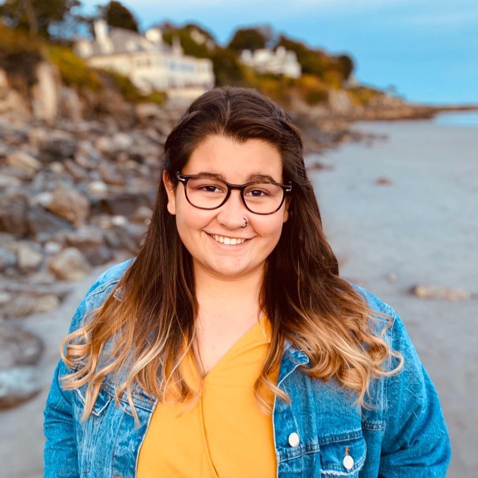 Woman with glasses and denim jacket smiling at the beach.