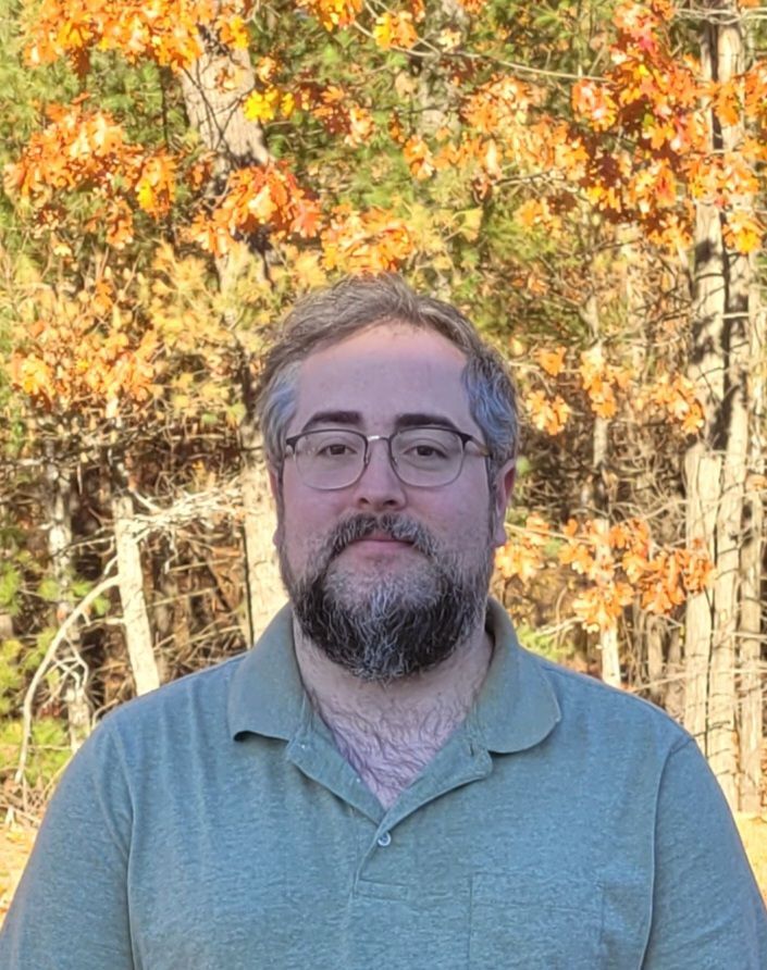 Man with a beard and glasses outdoors wearing a green polo shirt, with autumn foliage in the background.