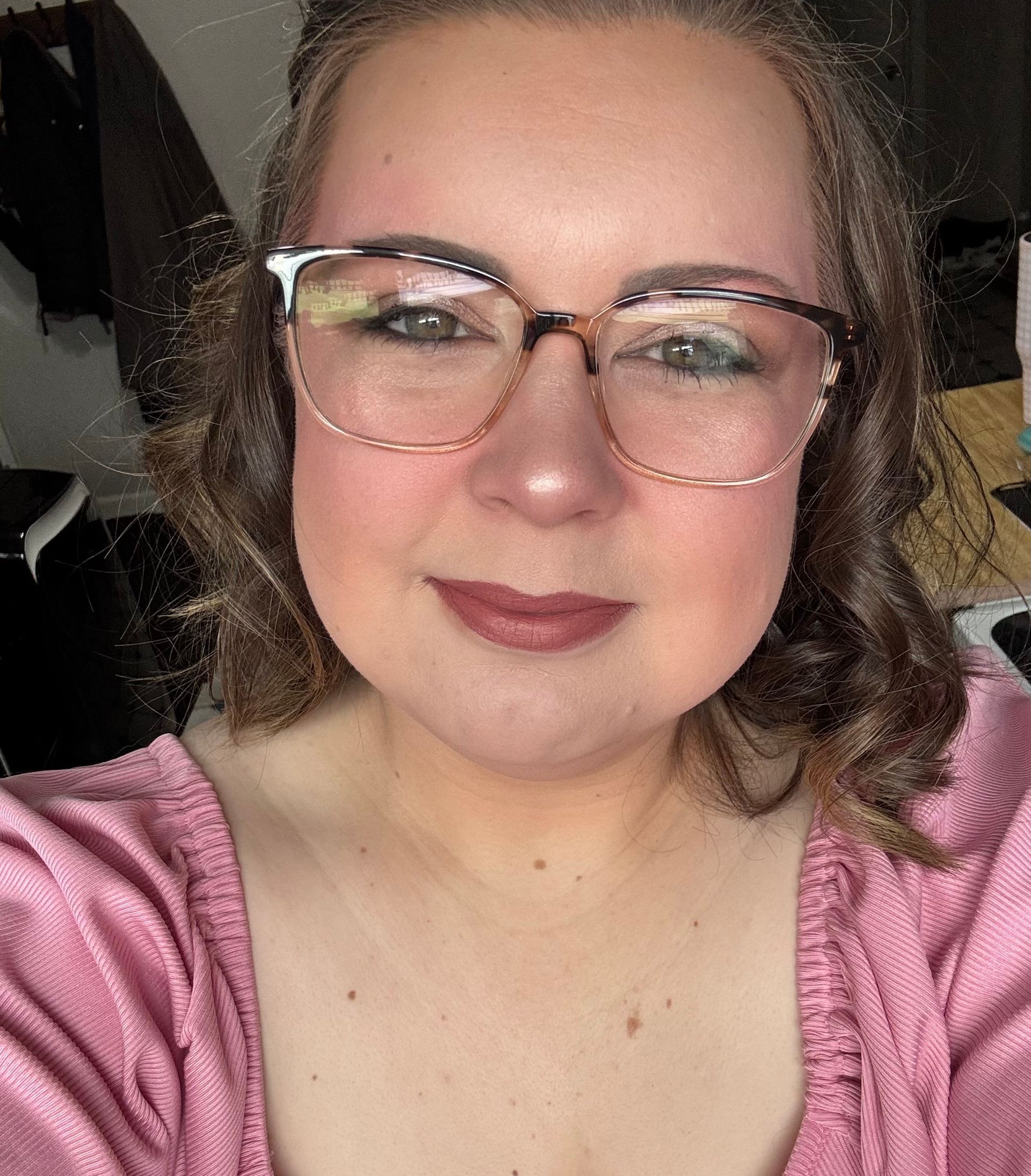 Woman with glasses, wearing a pink top, smiling. Brown hair and lipstick.