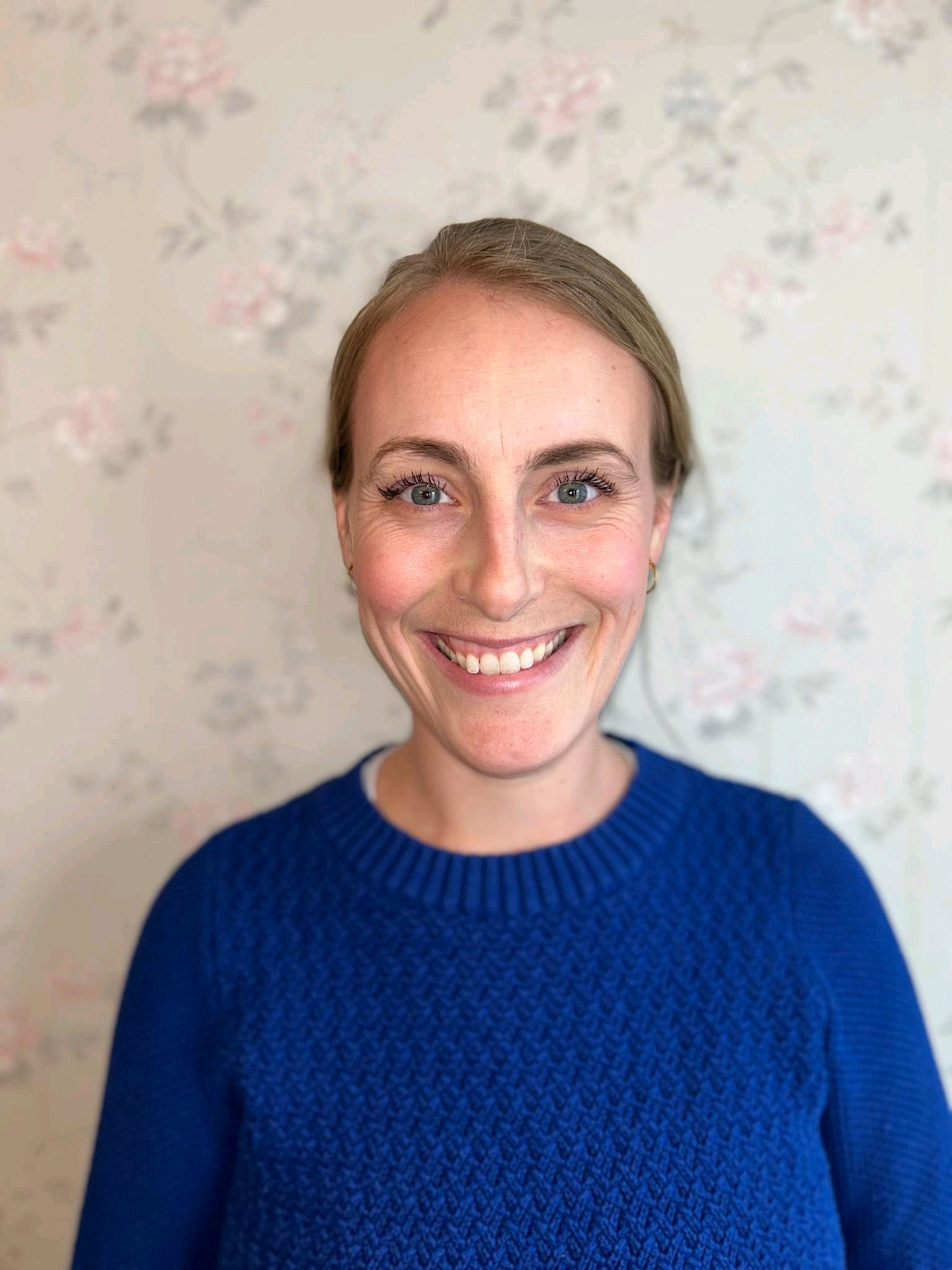 Woman with fair skin and blonde hair smiles in a blue sweater in front of a floral wallpaper.
