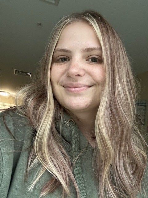 Woman with blonde hair and highlights smiles at the camera, wearing a green hoodie.