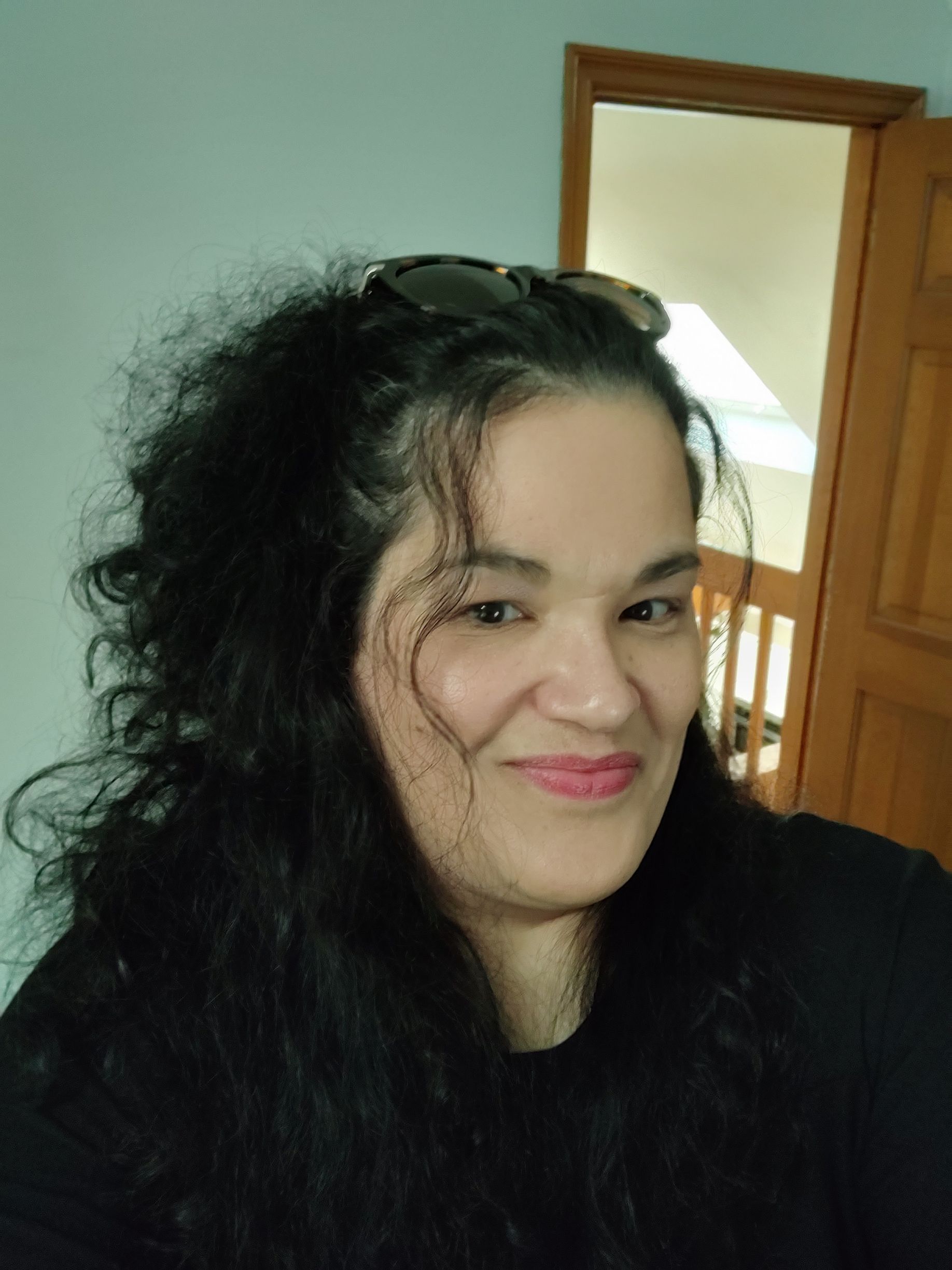 Woman with curly dark hair smiles, sunglasses on head, against a light blue wall.