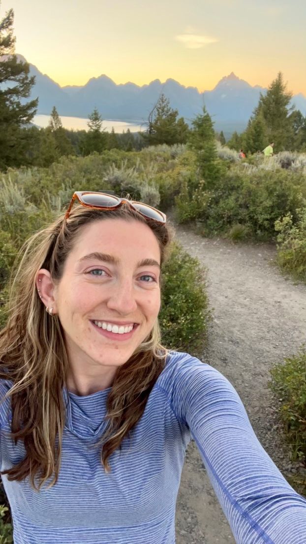 Woman smiling on a trail with mountains and sunset in the background.