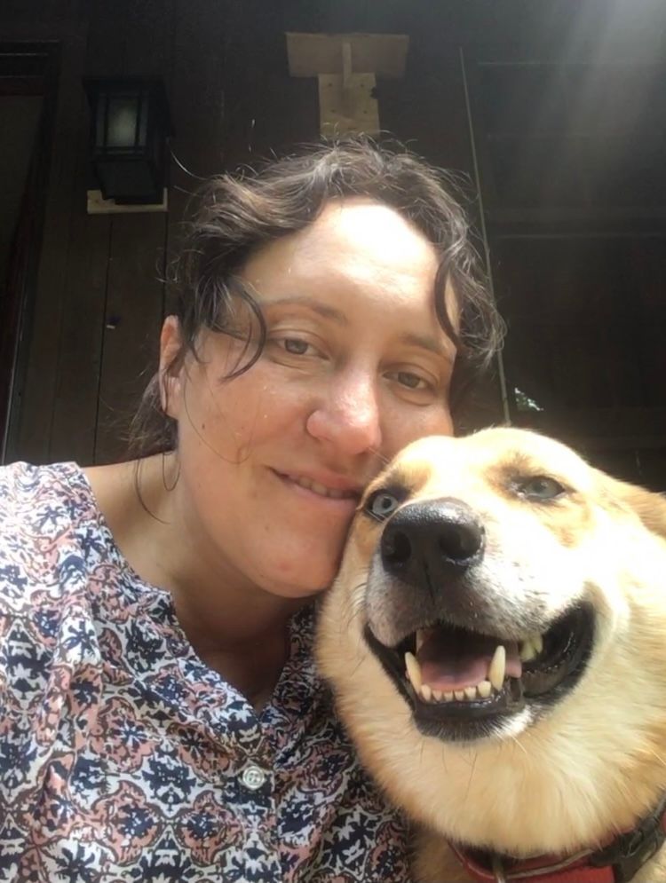 Woman and dog smiling close to the camera. Dog has one blue eye, tan and white fur. Woman has curly brown hair, floral shirt.
