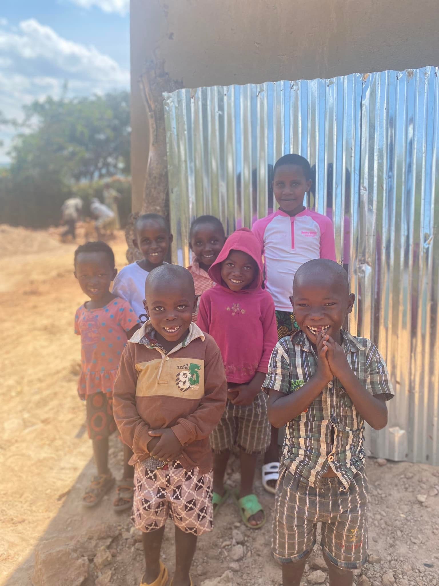 Group of smiling African children pose outdoors near a building with corrugated metal siding.
