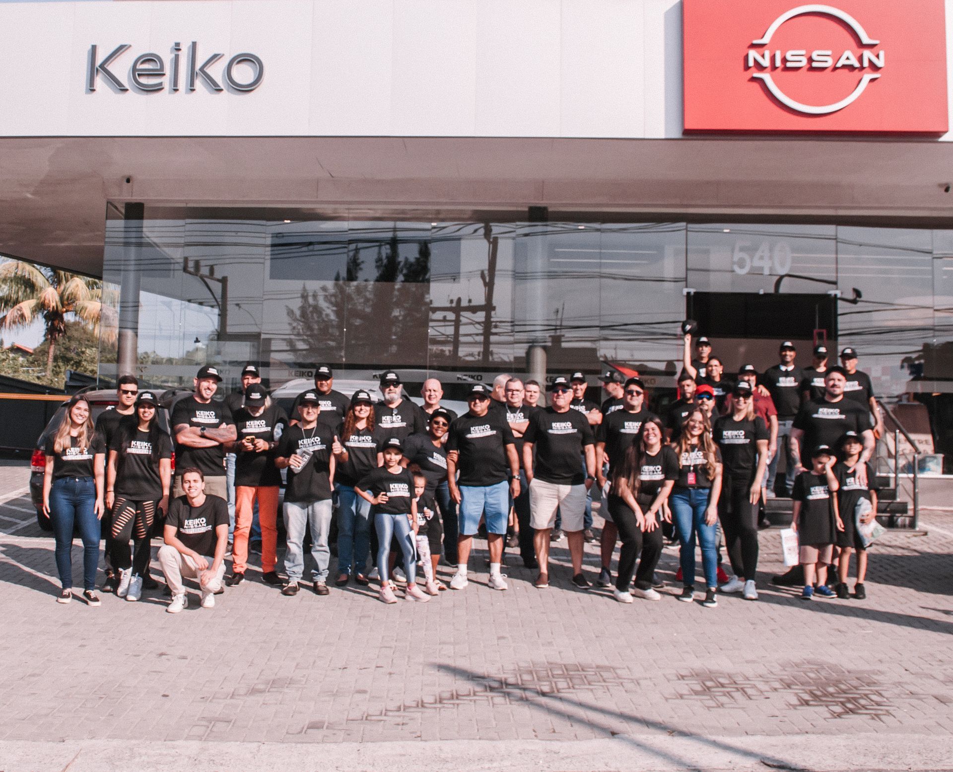 Grupo de pessoas vestindo camisetas pretas posando em frente à concessionária Keiko Nissan.