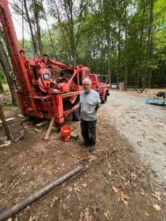 Man stands near a large red drilling machine in a wooded area. He gestures with his hands.