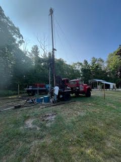 Red well-drilling truck with tall rig on a grassy lot, trees in the background, blue sky.