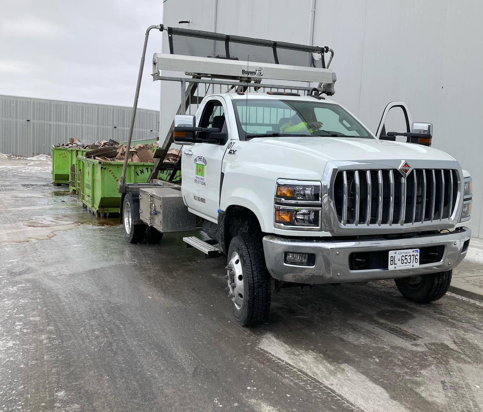 White truck with a lifting mechanism hauling green bins; outdoor setting.