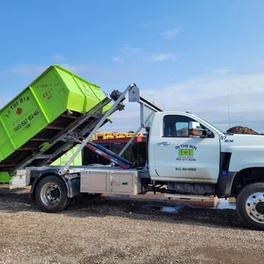 Green dumpster being lifted onto a white truck against a blue sky, for waste removal.
