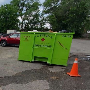 A green dumpster is sitting in a parking lot next to an orange cone.