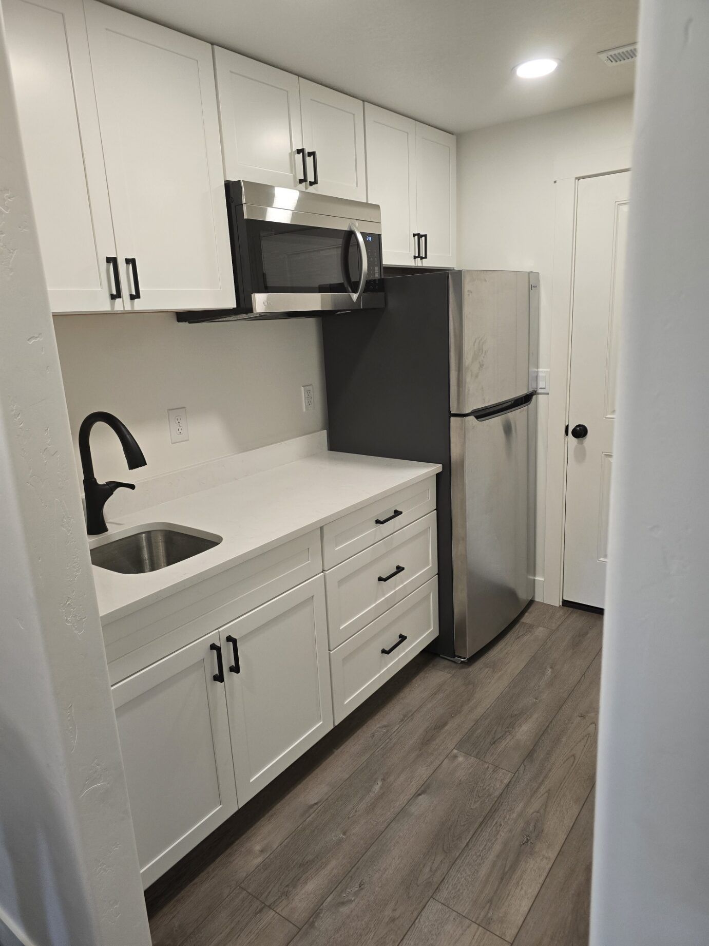 Small white kitchen with stainless appliances, sink, and wood-look floor.