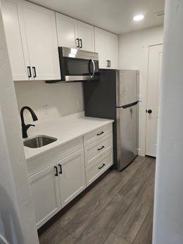 Small white kitchen with stainless appliances, sink, and wood-look floor.