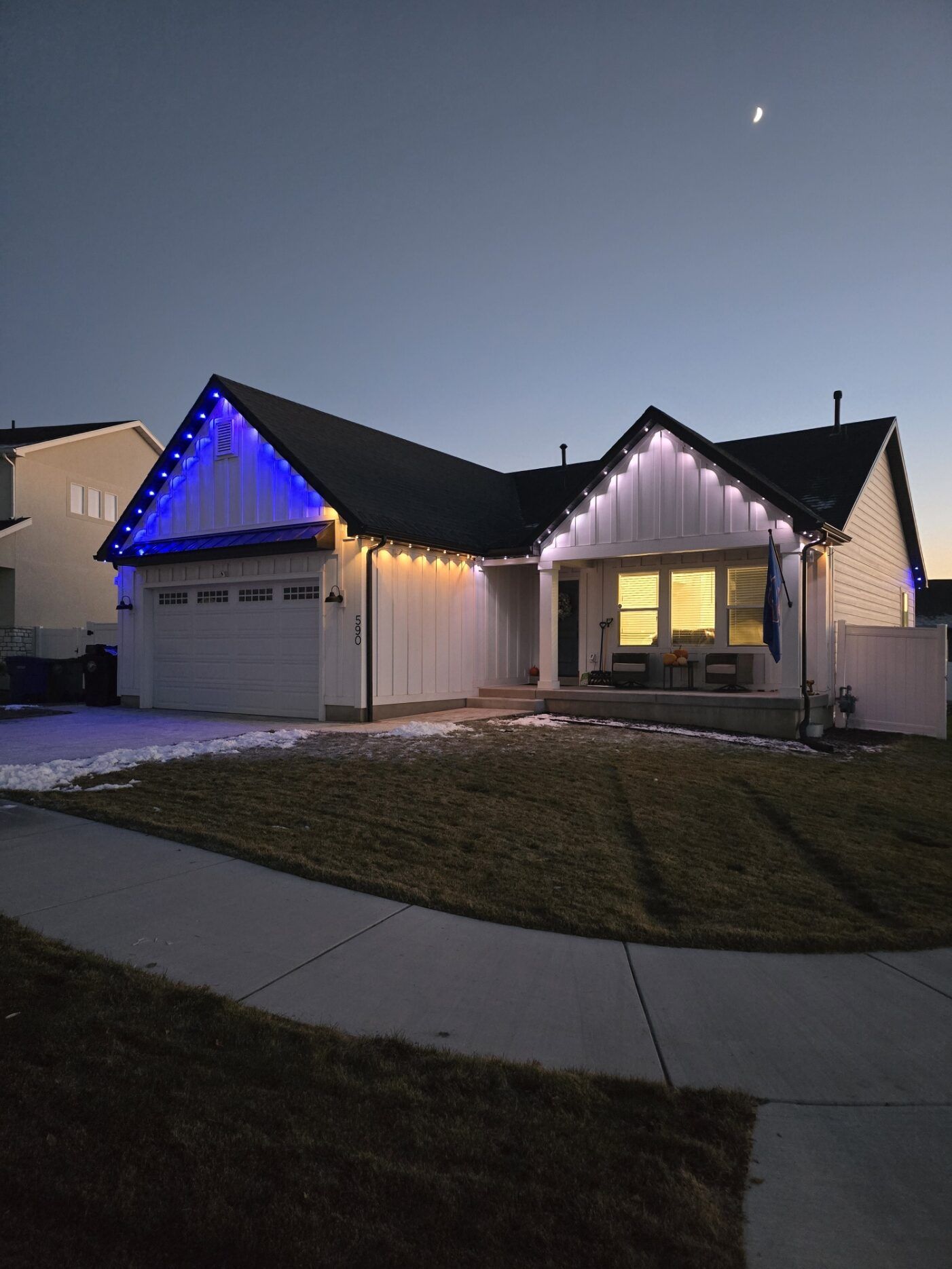 House at dusk with blue and white holiday lights along the roofline and a lit front window