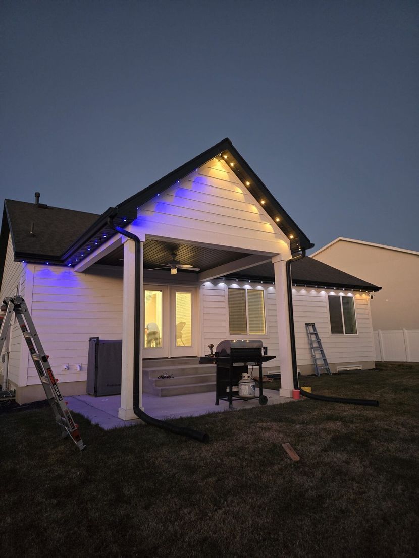Small white house at dusk with blue and warm lights on the porch and roofline.