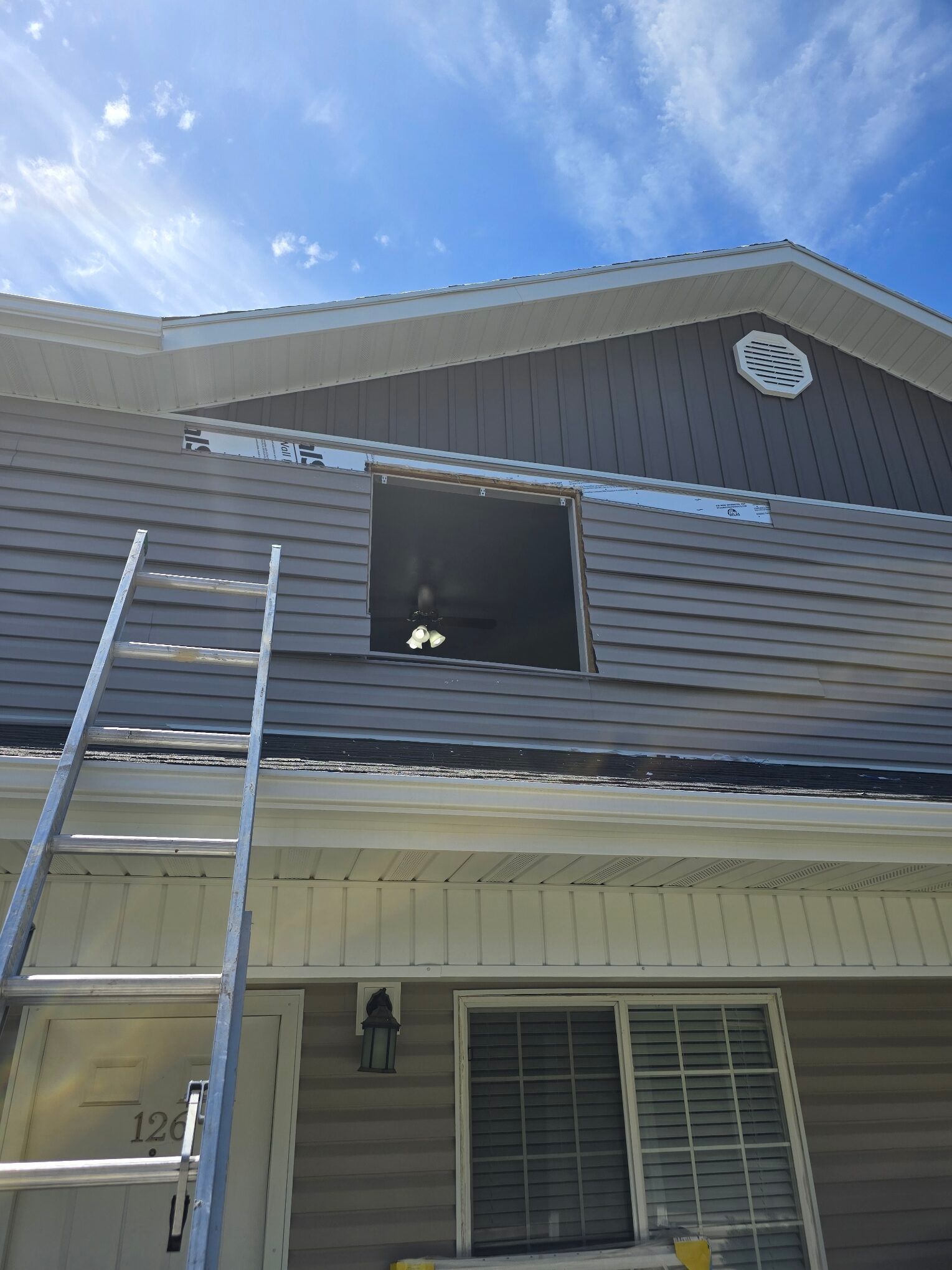 Exterior of a two-story house with gray siding, a ladder, and an open upper window under a blue sky