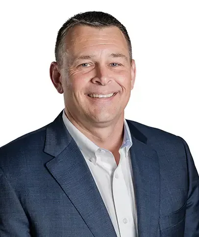 A person with short hair smiles, wearing a white collared shirt and a dark pinstriped blazer in an office setting.