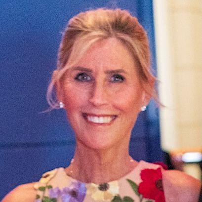 A person with long blonde hair and a friendly smile, centered in a close-up portrait against a dark, leather-like background.