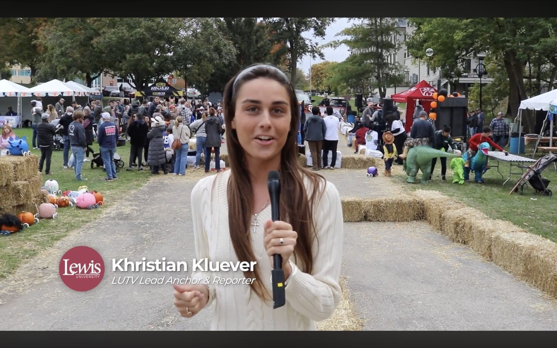 A reporter holds a microphone in a park during a fall festival with hay bales, pumpkins, and a crowd in the background.