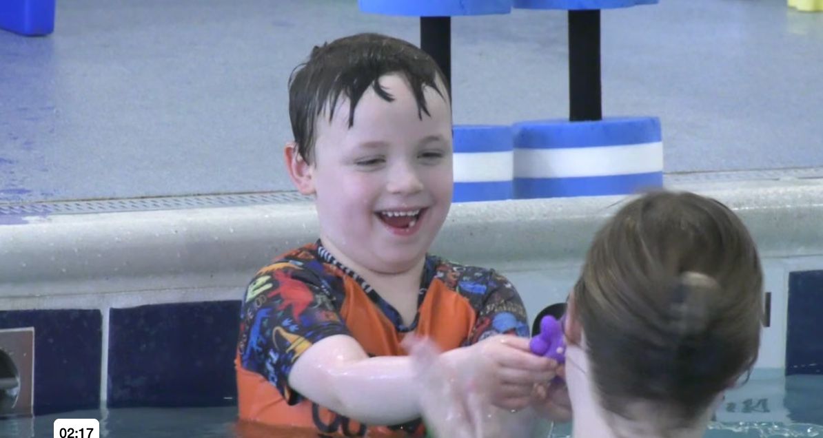 Child smiling in a pool, splashing water with another child nearby.