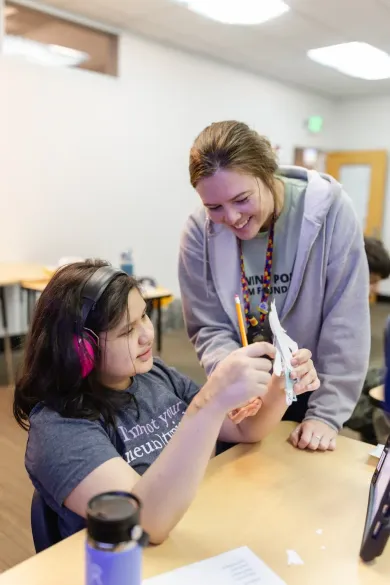 A smiling instructor works with a student wearing headphones, both focused on a small, white model held at a desk.