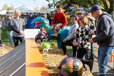 A group of people at an outdoor pumpkin race, with children interacting with a blue dinosaur prop and pumpkins on wheels.
