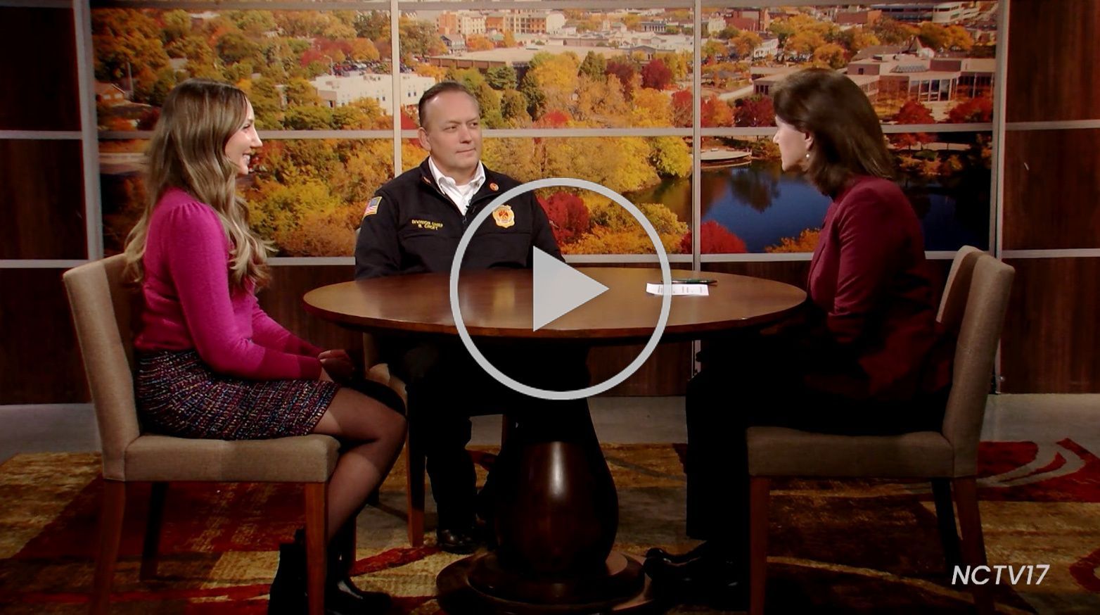 Two people sitting at a table with a moderator in a TV studio setting, with a fall-themed window display behind them.