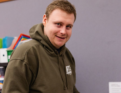 A smiling person with light brown hair wears an olive green hooded sweatshirt in an office with a grey wall and shelves.