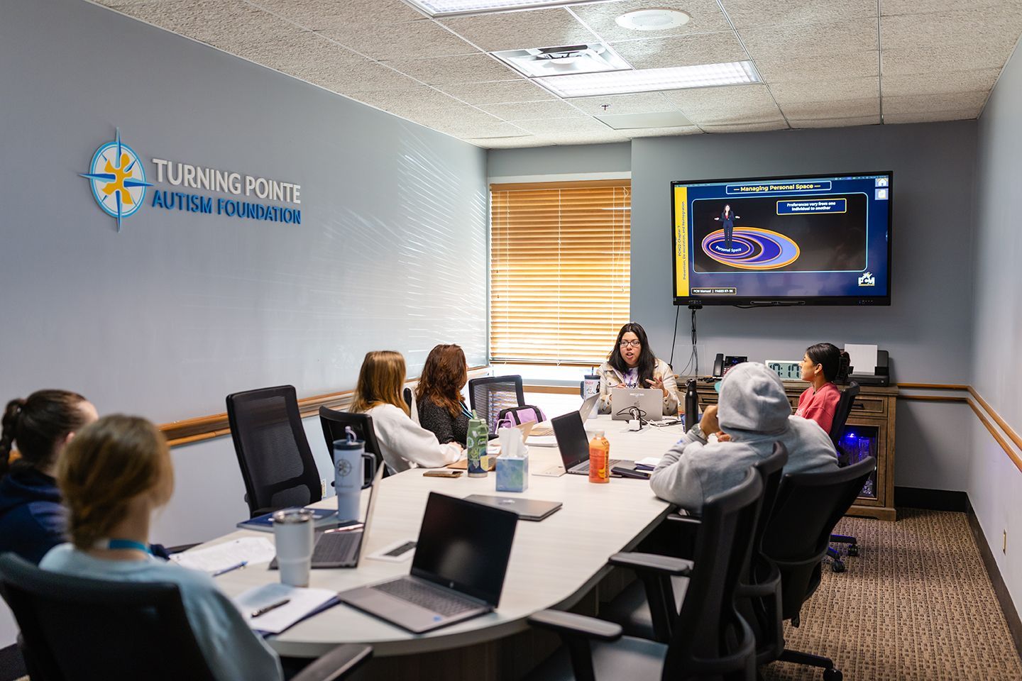 A group of people sitting around a conference table in an office, facing a monitor displaying a presentation.