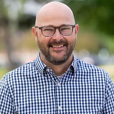 Smiling professional with a beard and glasses, wearing a blue and white checkered shirt against a soft, blurred background.