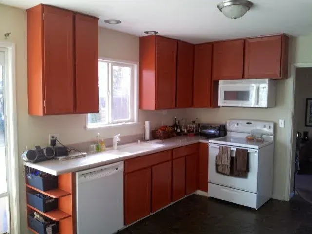 Orange kitchen cabinets and countertops in a room with a window, white appliances, and dark flooring.