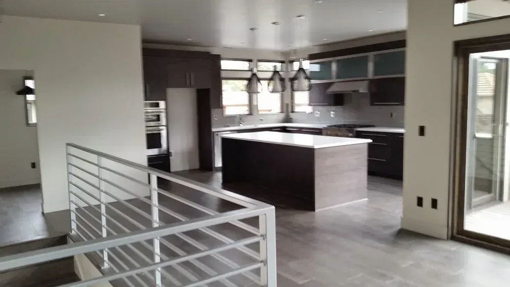 Modern kitchen with brown cabinetry, island, and tile flooring; adjacent staircase with metal railing.