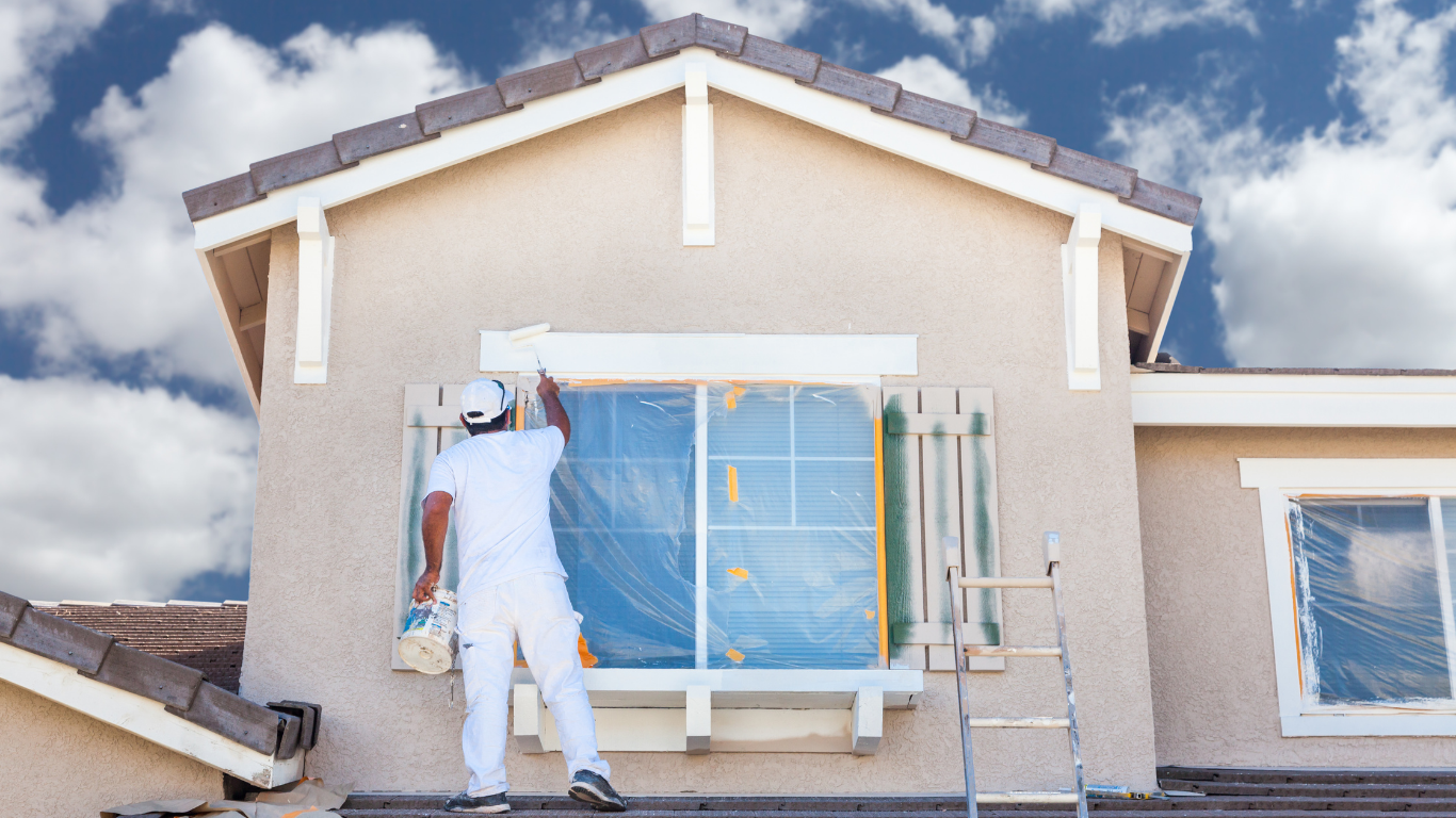 Painter in white overalls on a ladder, painting trim of a house with covered windows against a blue sky.