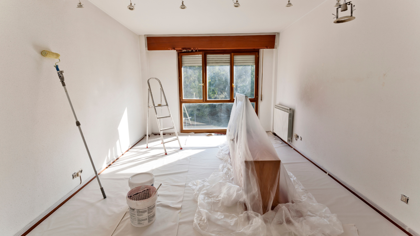 Room being painted, with roller, ladder, window, covered furniture, and drop cloths. White walls.