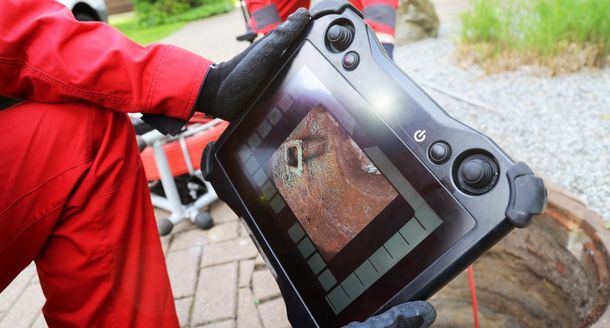 Gloved person holding a black handheld drone controller with live camera view on screen outdoors