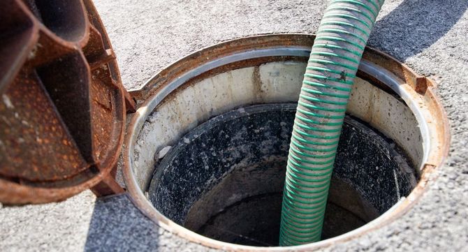 Green hose inserted into an open underground manhole with a rusty metal cover nearby.