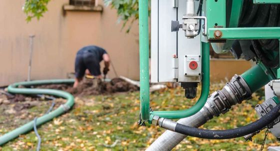 Worker inspecting green pipes and machinery outdoors with hose on leaf-covered ground
