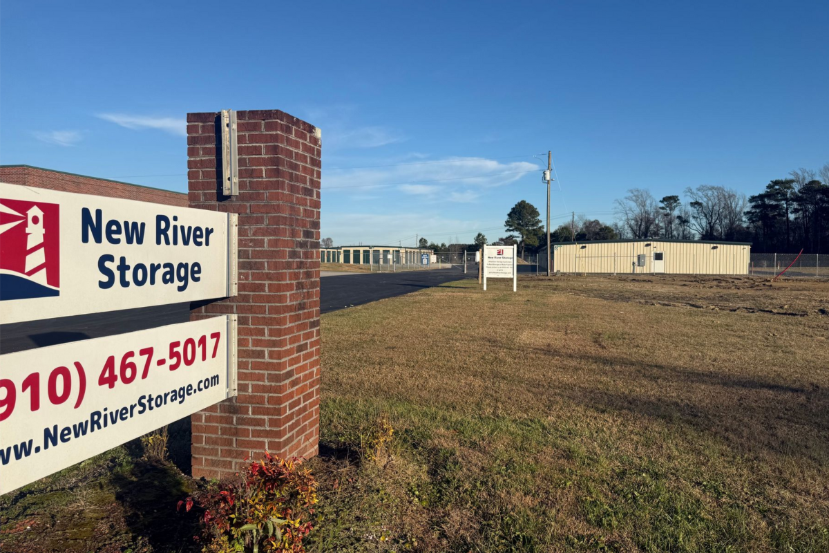A brick sign for New River Storage in Jacksonville, NC. Buildings in the background with Climate Controlled Self Storage.