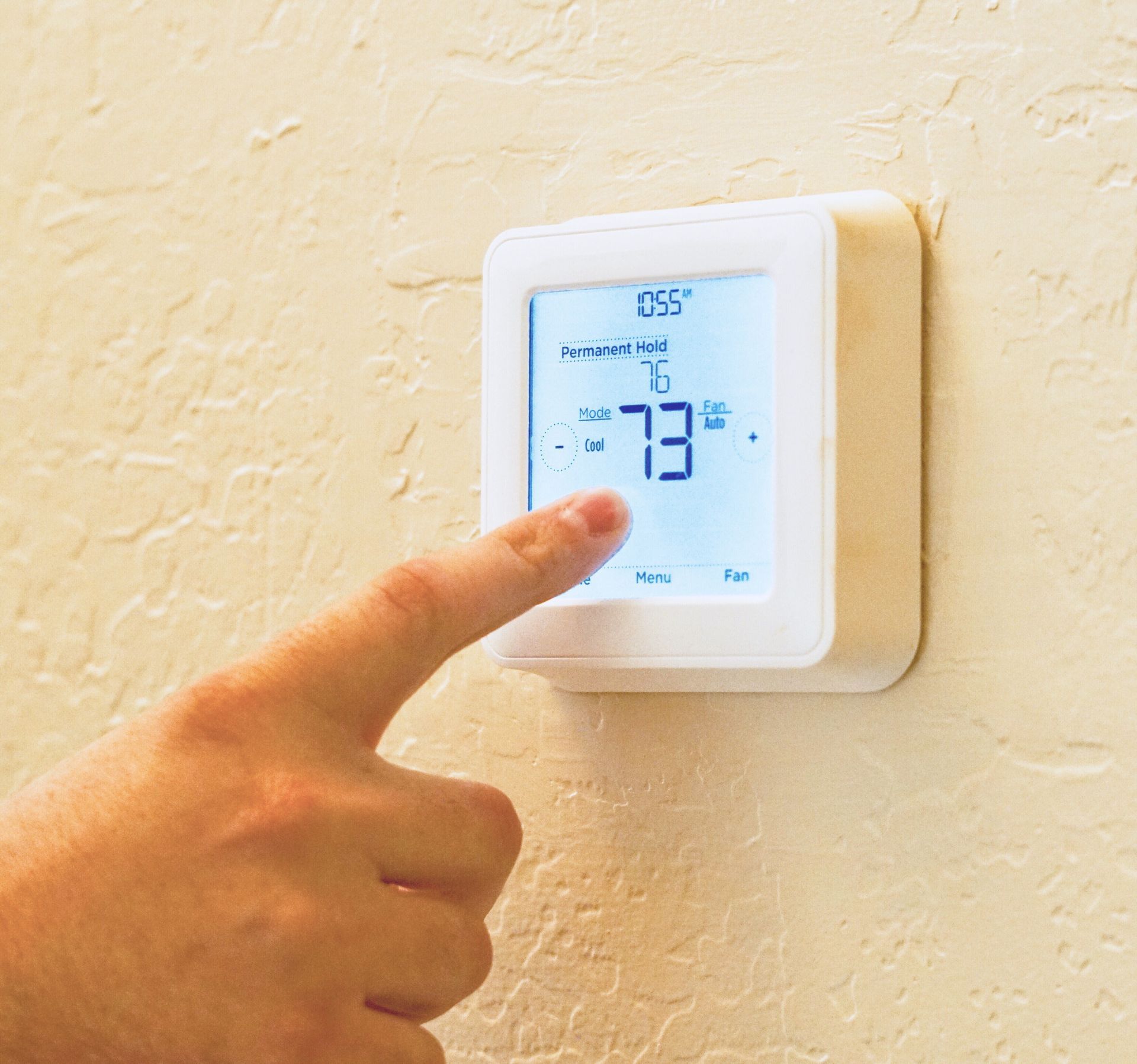 Hand adjusting a white wall thermostat showing 73°F on a beige textured wall