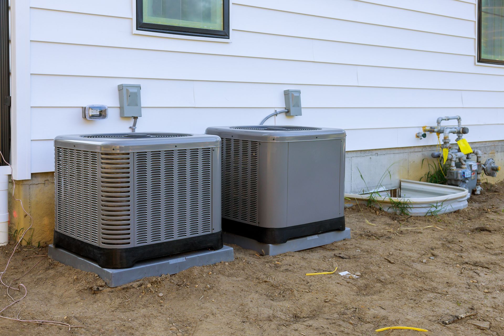Two outdoor HVAC units beside a house with white siding and a small flower bed.