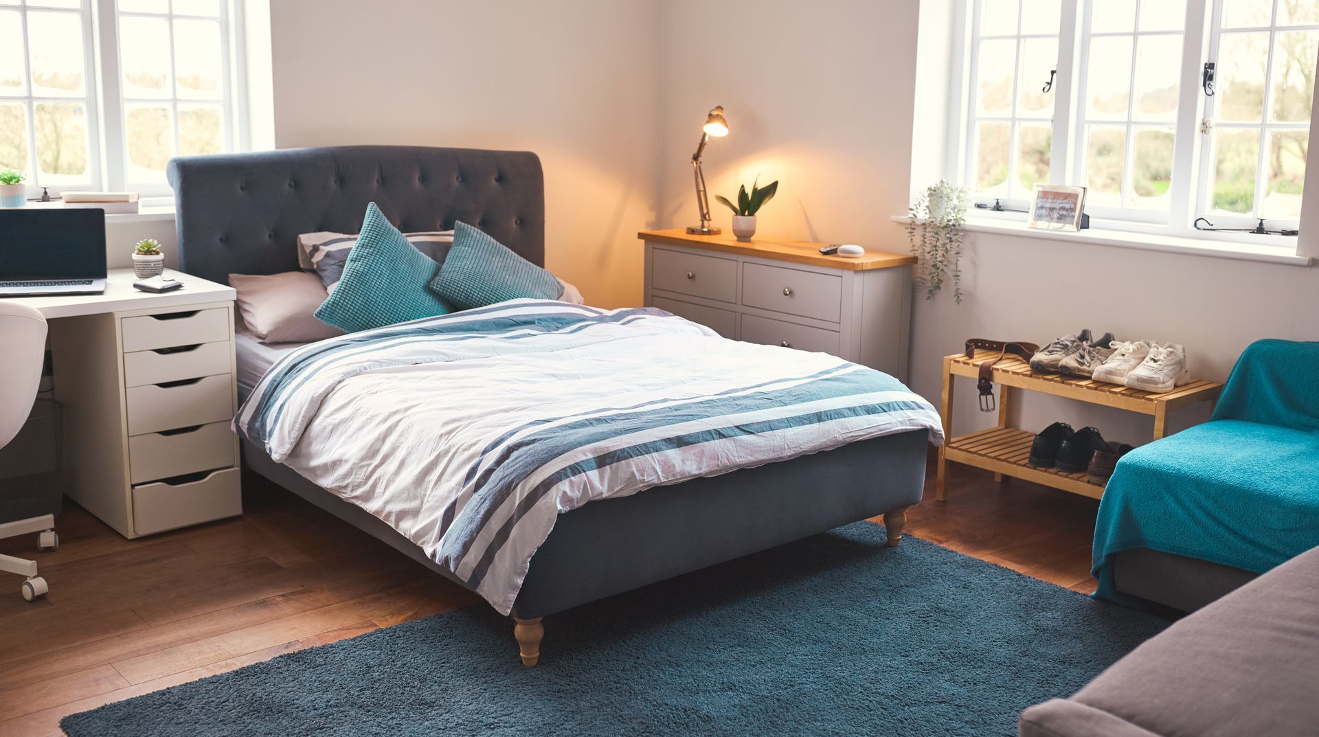 Bright bedroom with a gray bed, teal accents, white desk, and warm lamp beside a window