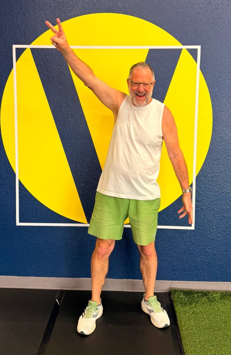A man in a white tank top and green shorts stands on a mat, raising his arms in a triumphant pose.