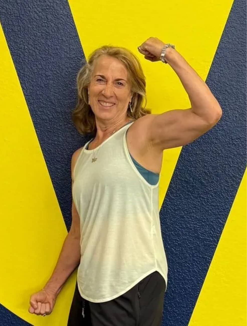Woman in a sleeveless top flexes her arm and smiles confidently. She stands against a bold blue and yellow background, exuding strength and positivity.